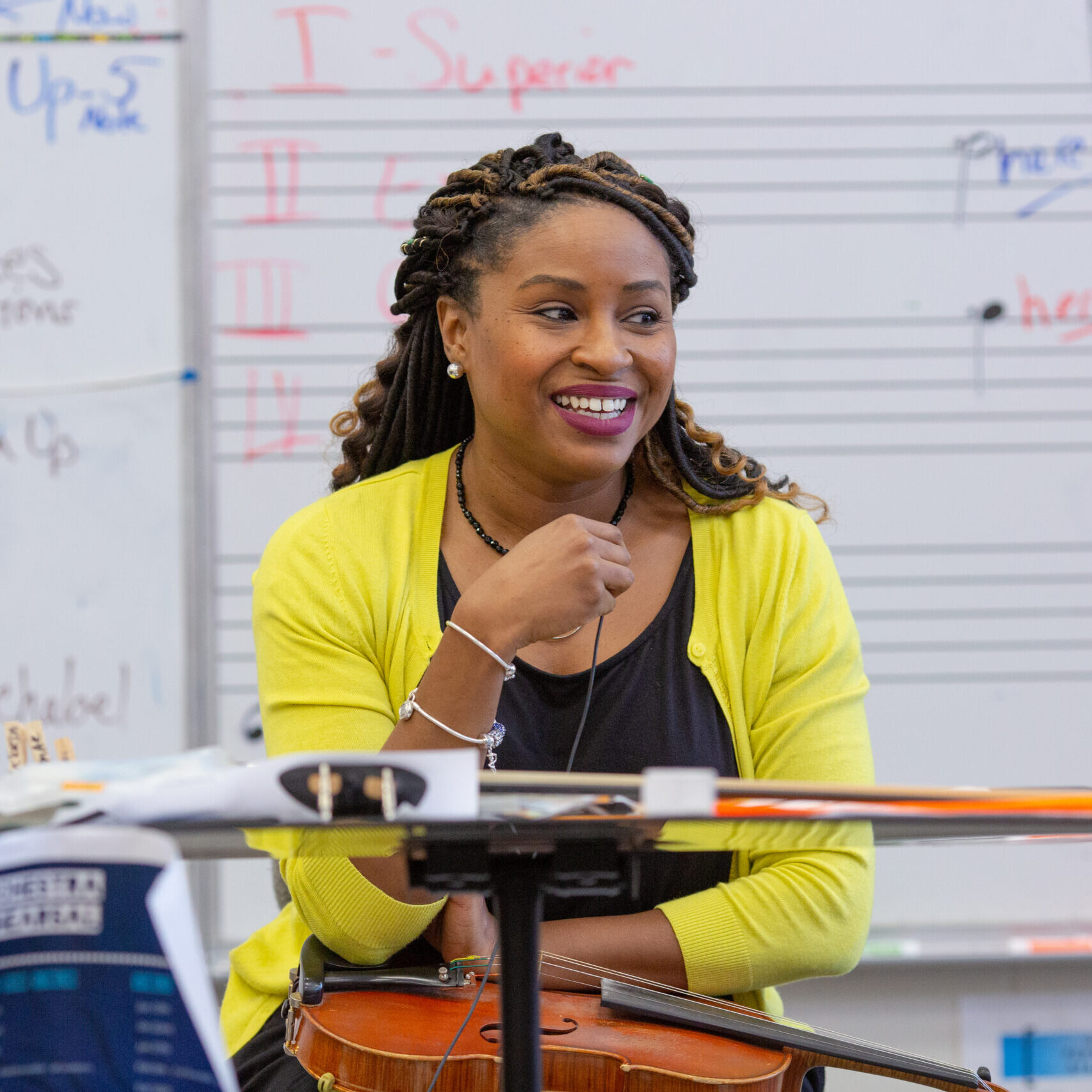 A young black woman sits at the front of a band classroom smiling at her students.